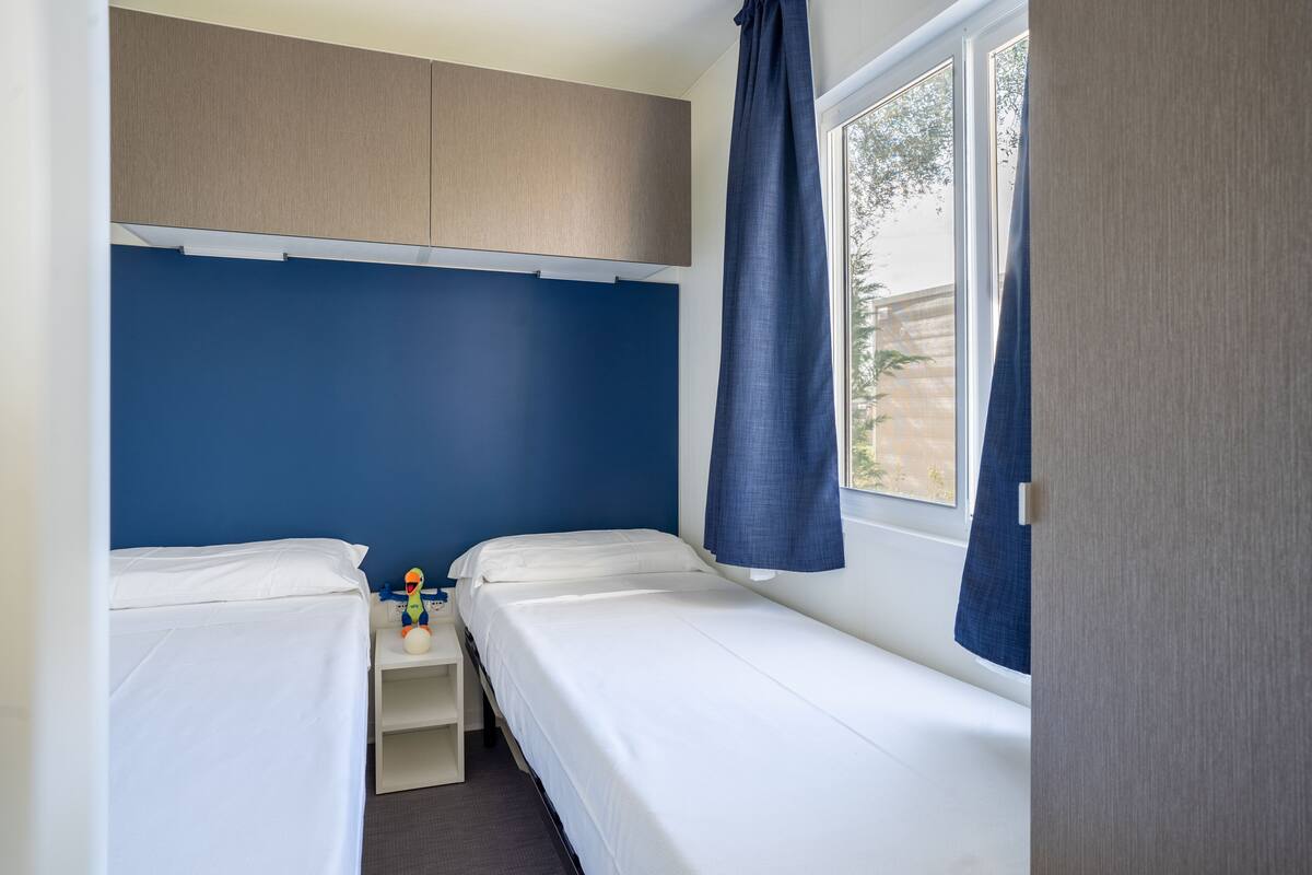 A small, single-room hotel room with two beds, a window with curtains, and a nightstand with a lamp and a toy.