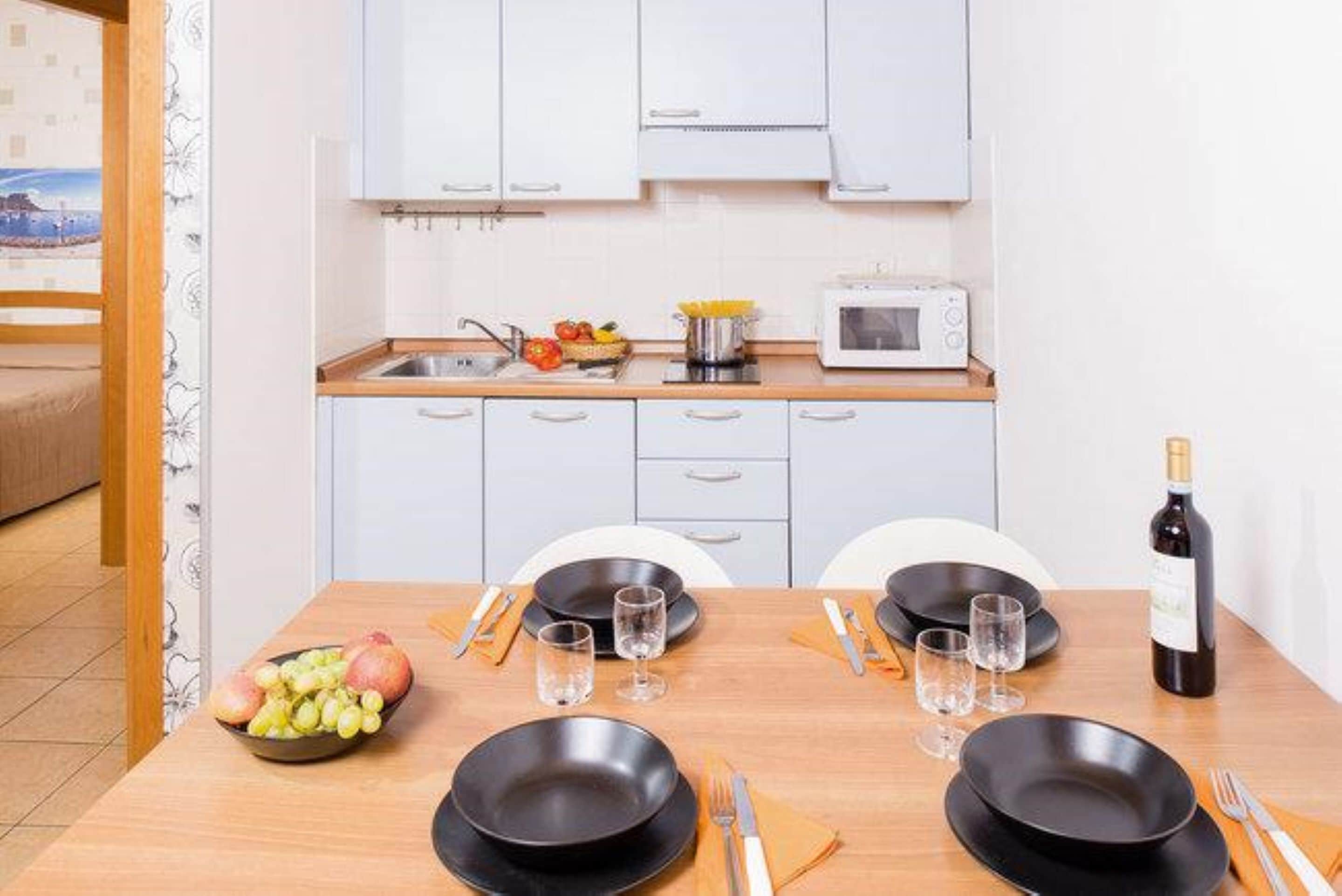 Una mesa de comedor dispuesta con platos negros, vasos y cubiertos, un frutero y una botella de vino.
