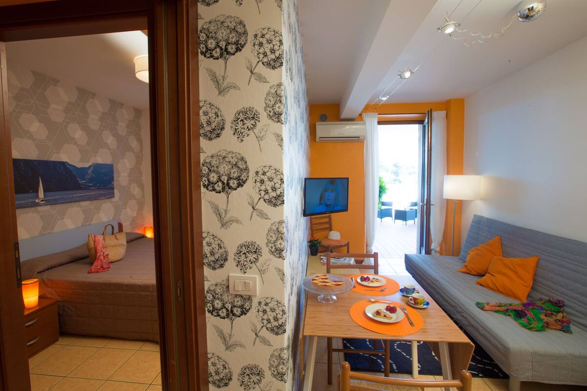 A cozy room with a bed, a dining table with orange placemats, a sofa with orange pillows, and a TV on the wall.