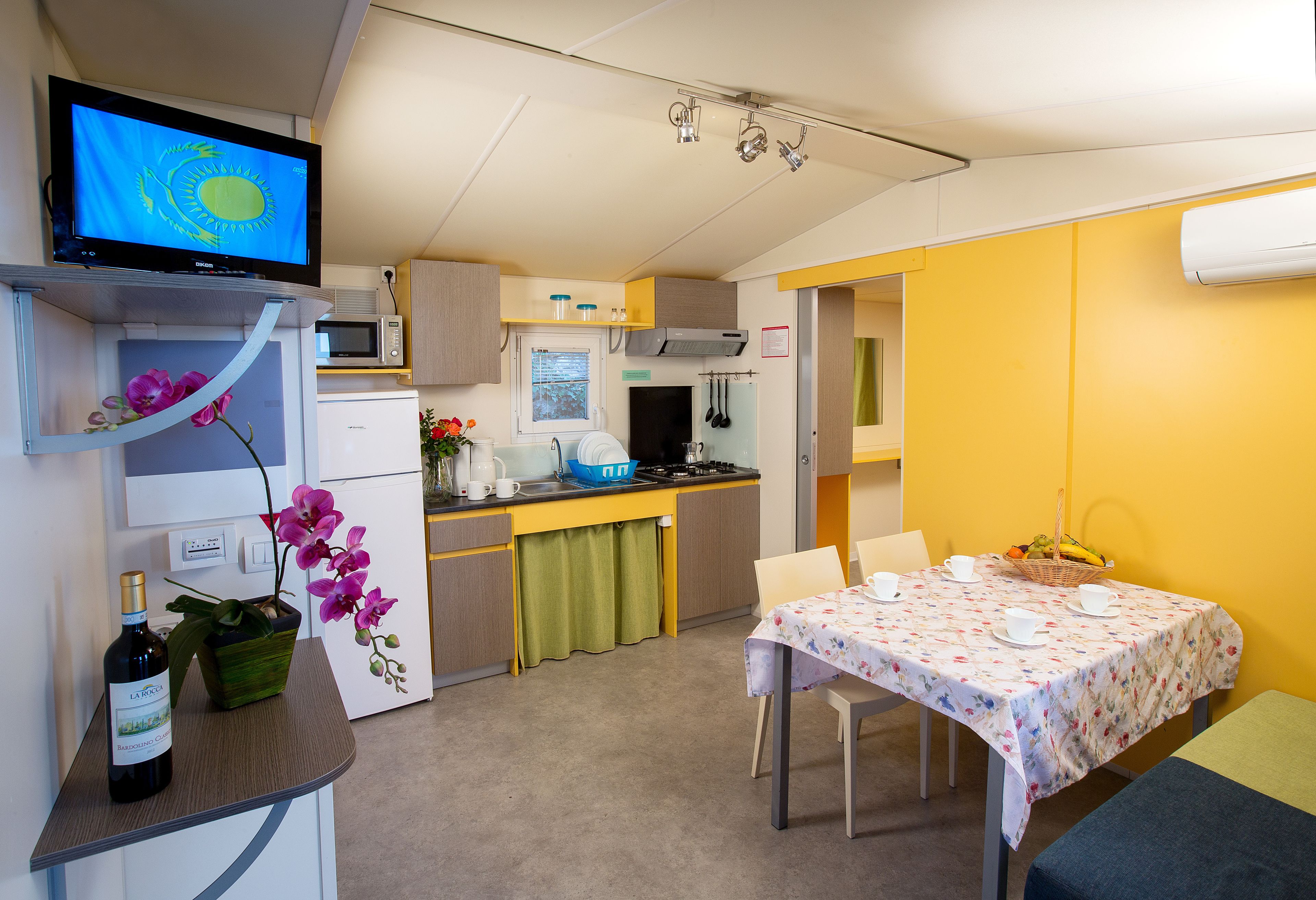 Une cuisine compacte équipée d’un four à micro-ondes, d’une cuisinière et d’un évier. On y trouve une table à manger avec des chaises, et un petit téléviseur fixé au mur au-dessus de la table.