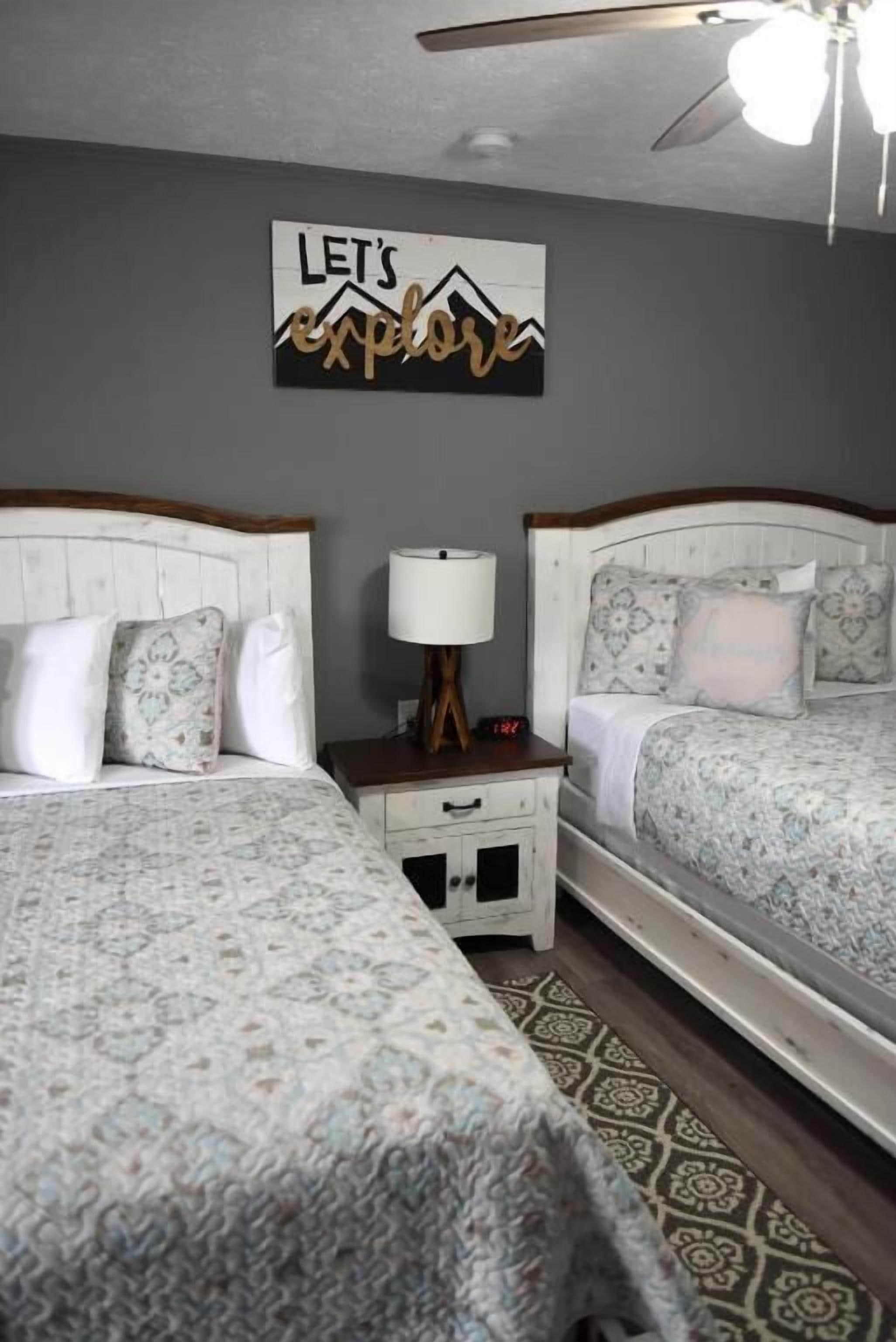A bedroom with two beds, a nightstand with a lamp, a wall clock, and a 'Let's explore' sign.