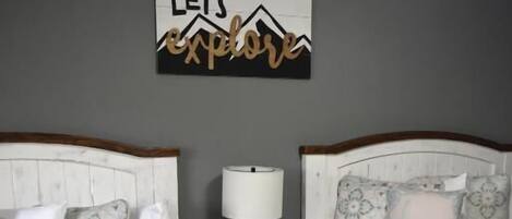 A bedroom with two beds, a nightstand with a lamp, a wall clock, and a 'Let's explore' sign.