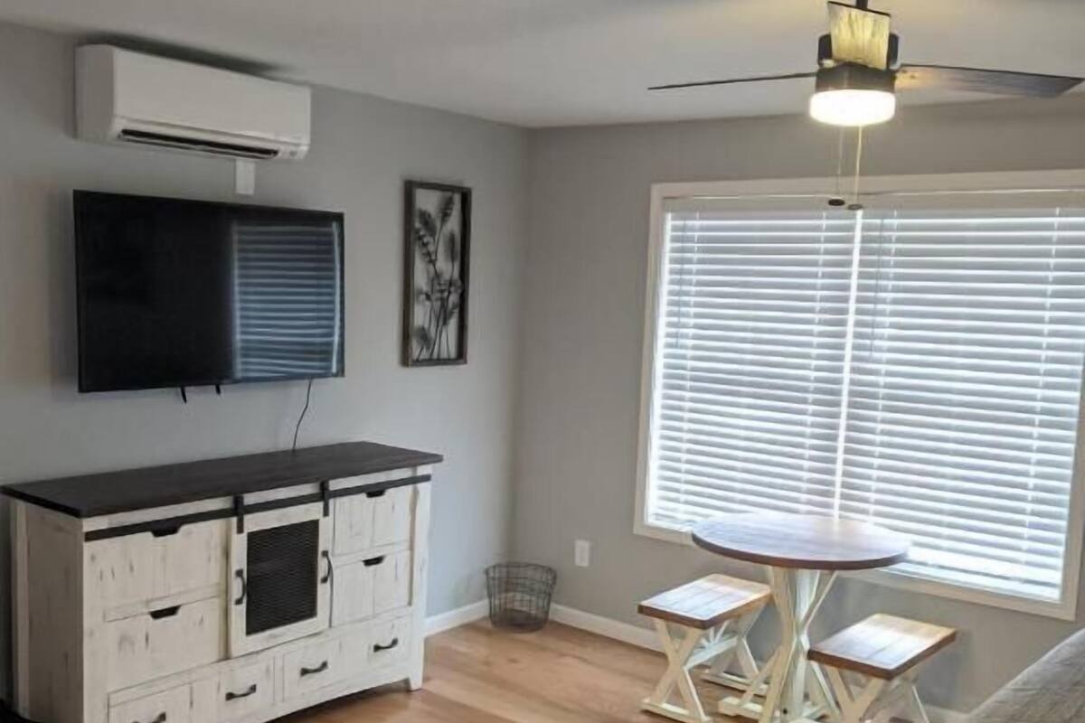 A room with a ceiling fan, a large window with blinds, a white cabinet with drawers, and a small table with two stools.