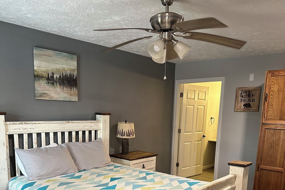 A bedroom with a wooden bed, a ceiling fan, a nightstand with a lamp, a door, and a wall with a picture and a sign.