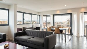A modern living room with a grey sofa, dining table, chairs, and large windows offering a city view. - The Originals Residence, Appart'Hôtel Hévéa, Valence (Valence)