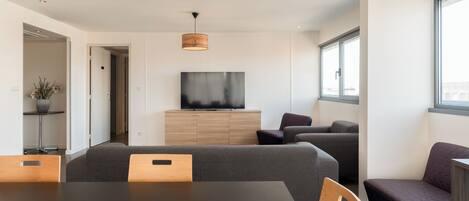 Una sala de estar moderna con una mesa de comedor, sofás, un televisor sobre un mueble de madera y ventanas amplias.