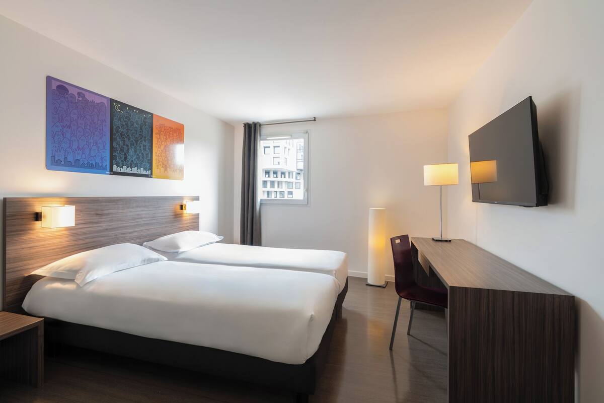 A hotel room with two beds, a desk, a chair, a TV, and artwork on the wall.