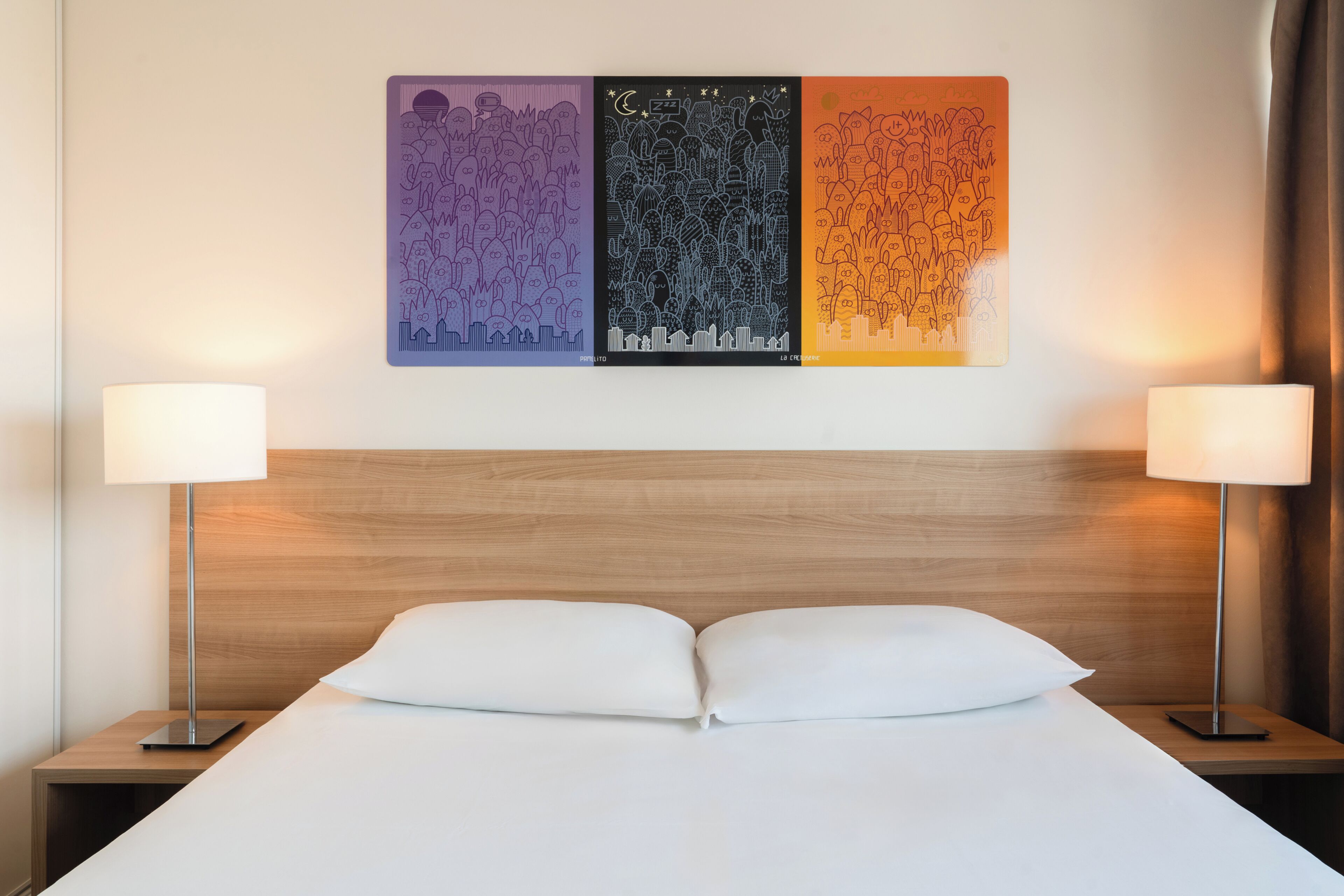 A hotel room with a bed, two bedside lamps, and three wall art pieces.