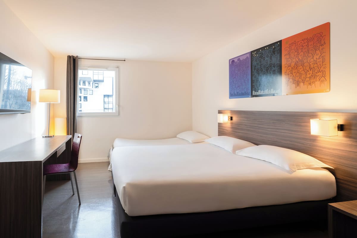 A hotel room with a bed, desk, chair, lamp, and artwork on the wall.