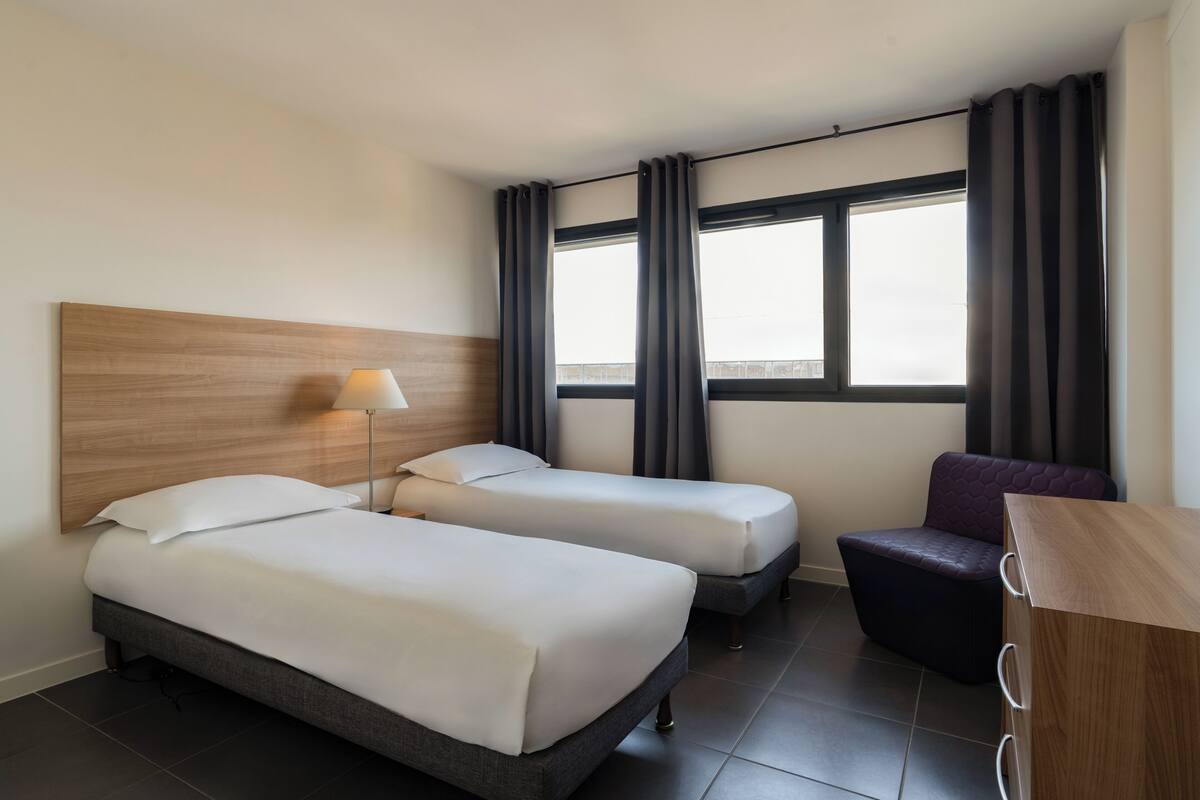 A hotel room with two beds, a desk, a chair, a lamp, and a window with curtains.