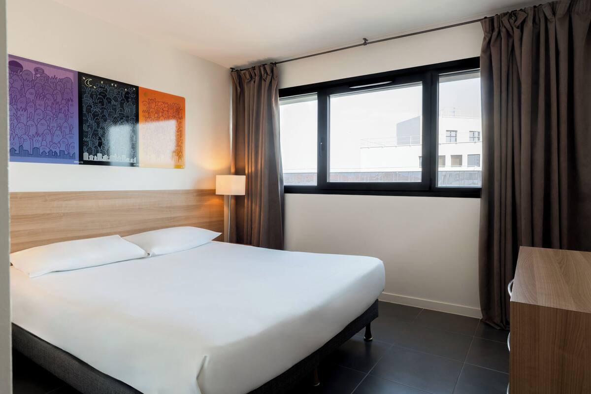 A hotel room with a bed, a window with curtains, a wall with artwork, and a bedside table.