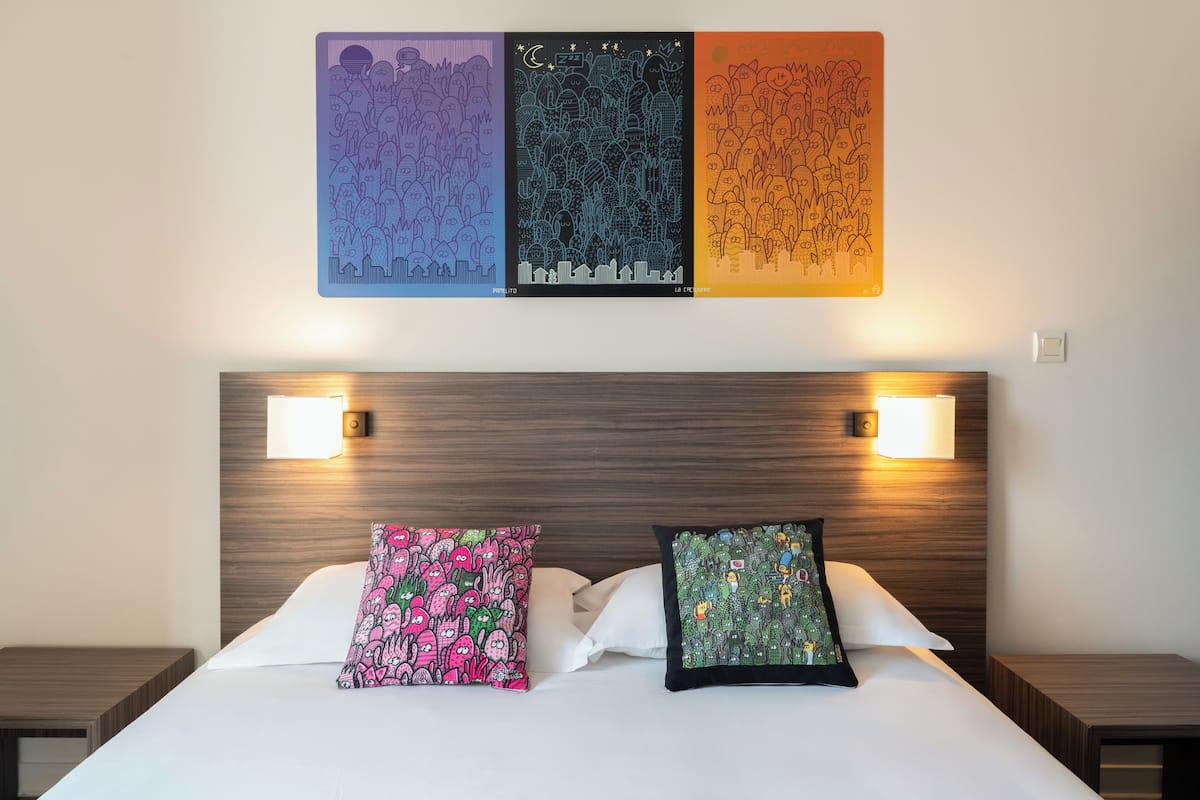 A hotel room with a bed, two pillows, a nightstand, a wall-mounted artwork, and two wall-mounted lamps.