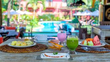 A dining table with plates of food, a green drink, and a dessert, set against a view of a pool and outdoor seating.