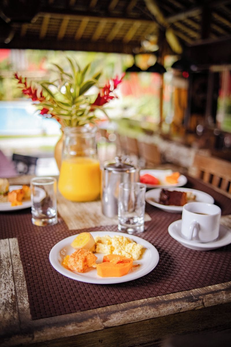 A dining table with breakfast items, a glass of orange juice, and a vase with flowers.