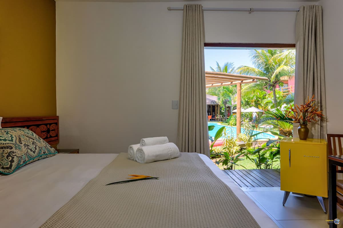 Camera d'albergo con un letto, un tavolino, un armadietto giallo e vista su piscina e verdeggiante vegetazione all'esterno.