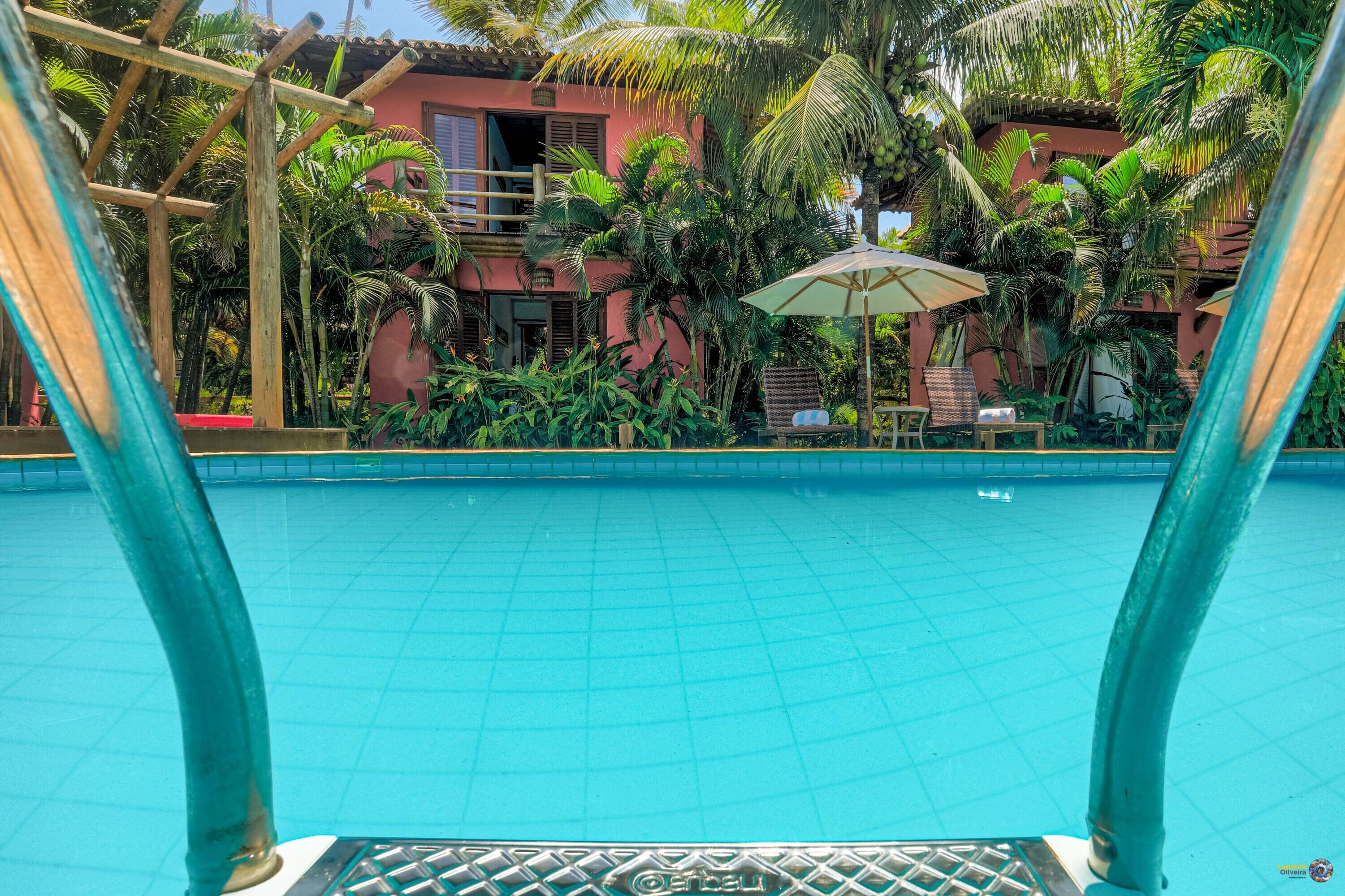 A swimming pool with a tiled floor, surrounded by lush greenery and a building with a red facade.