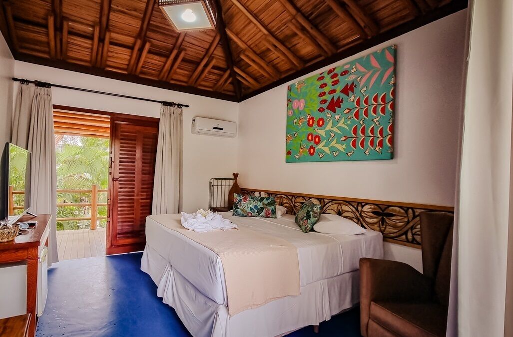 A bedroom with a wooden ceiling, a bed with white linens, a colorful painting on the wall, and a view of greenery outside.