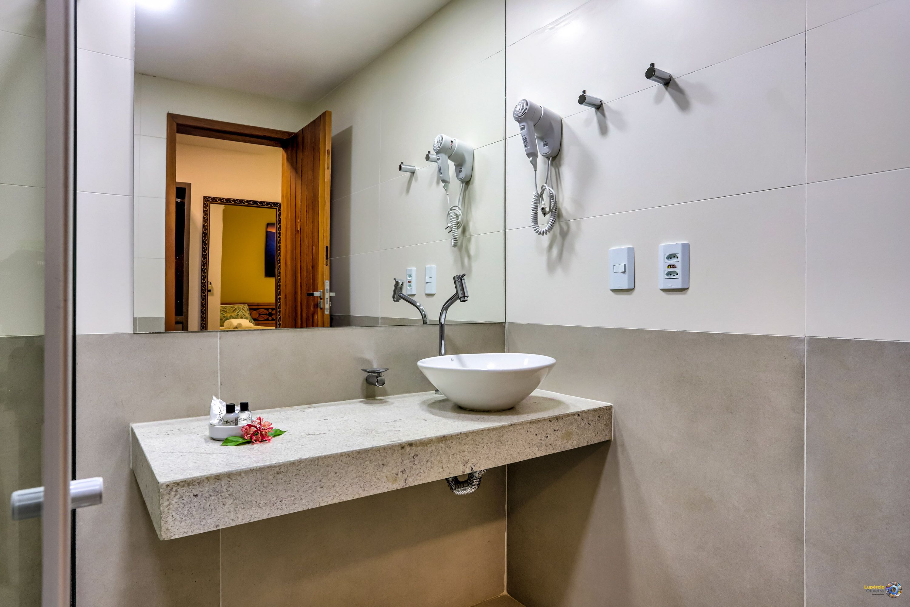 Een badkamer met een grote spiegel, een witte wastafel met kom en meerdere haardrogen op de muur.
