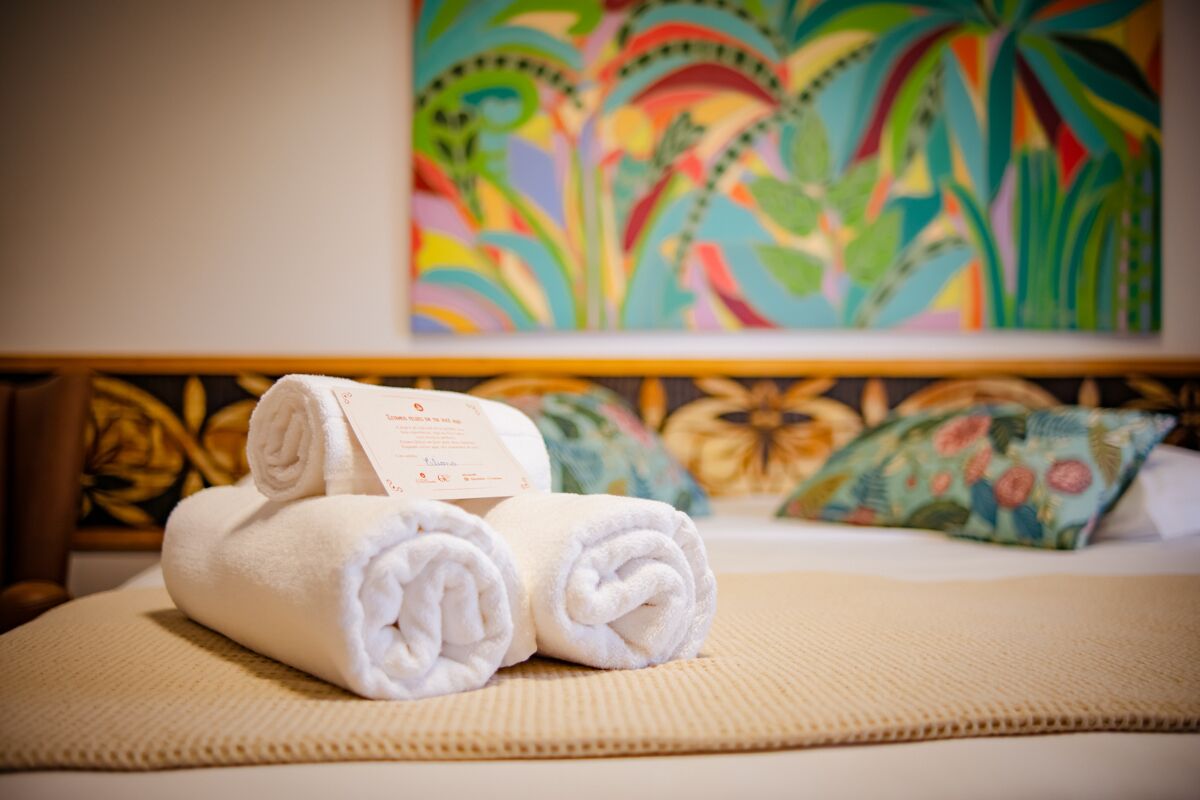 Una cama bien hecha con toallas blancas enrolladas y una tarjeta, un cuadro abstracto de colores en la pared y una almohada floral sobre la cama.