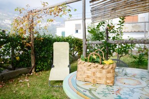 A wicker basket with lemons on a table with a floral tablecloth, a chair, and a view of a building and greenery.