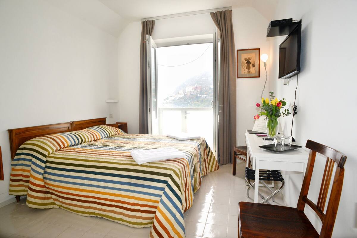 A hotel room with a bed, a desk with a chair, a television, and a window with curtains.
