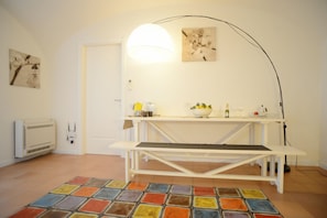 A room with a white table, a bench, a colorful tiled floor, a white radiator, and a wall-mounted lamp.