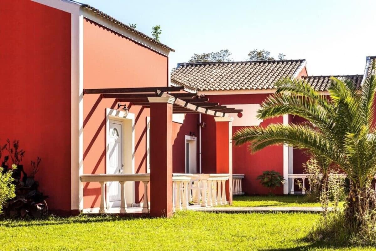 A red building with a white balcony, a palm tree, and a tiled roof.