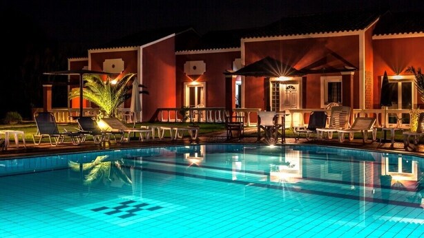 Una piscina con sillas de descanso y sombrillas, rodeada de edificios con iluminación cálida.