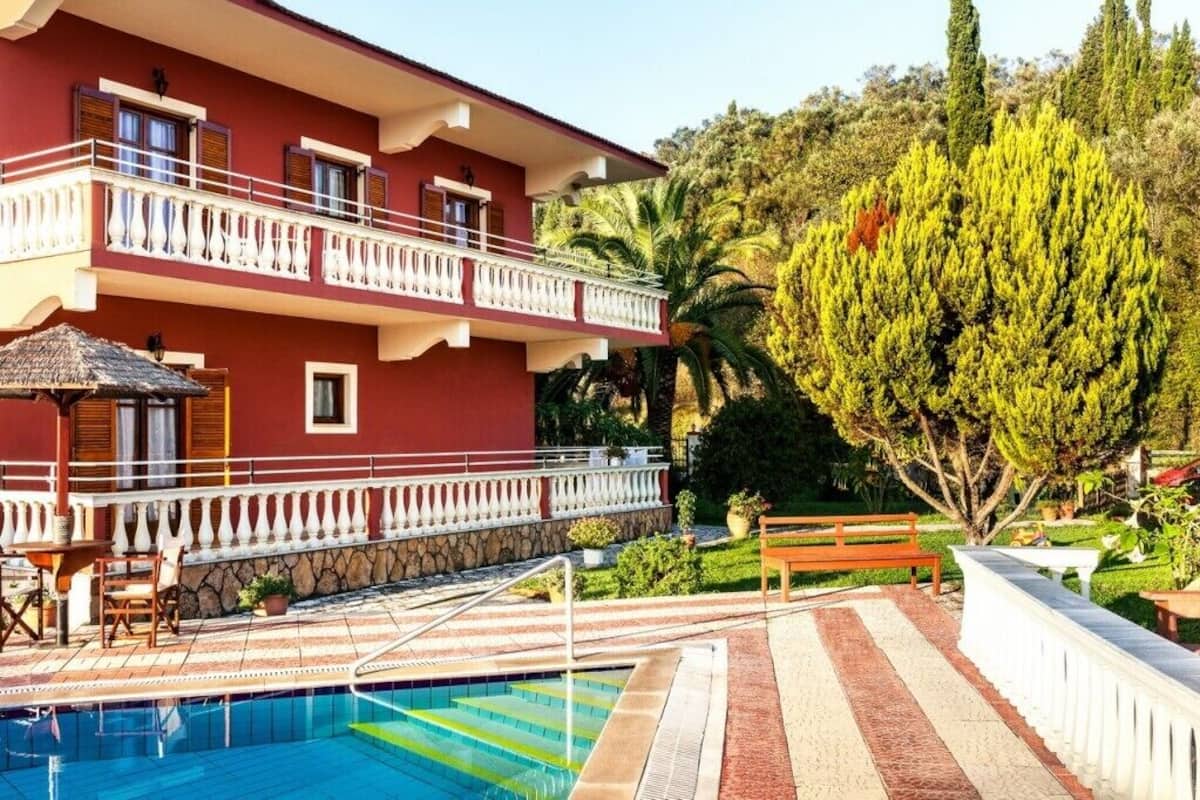 A hotel with a swimming pool, a balcony with white railings, and a large tree in the background.
