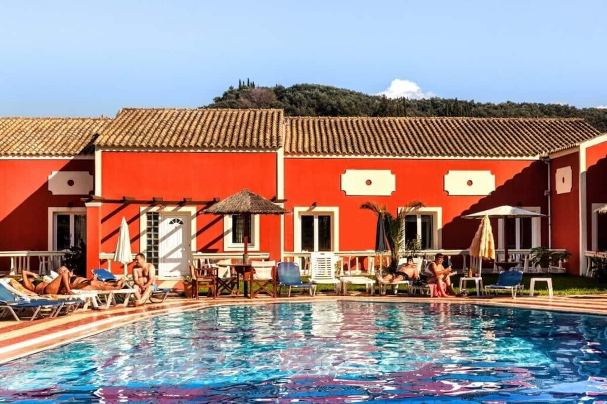 A swimming pool area with lounge chairs and umbrellas, a red building with a tiled roof, and a view of a hill in the background.