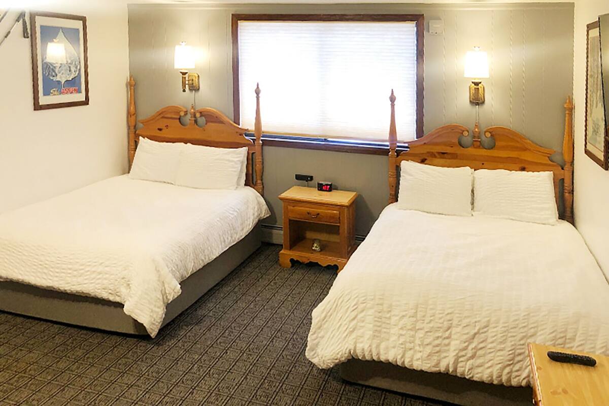 A hotel room with two single beds, a small wooden nightstand, a clock, and a window with blinds.