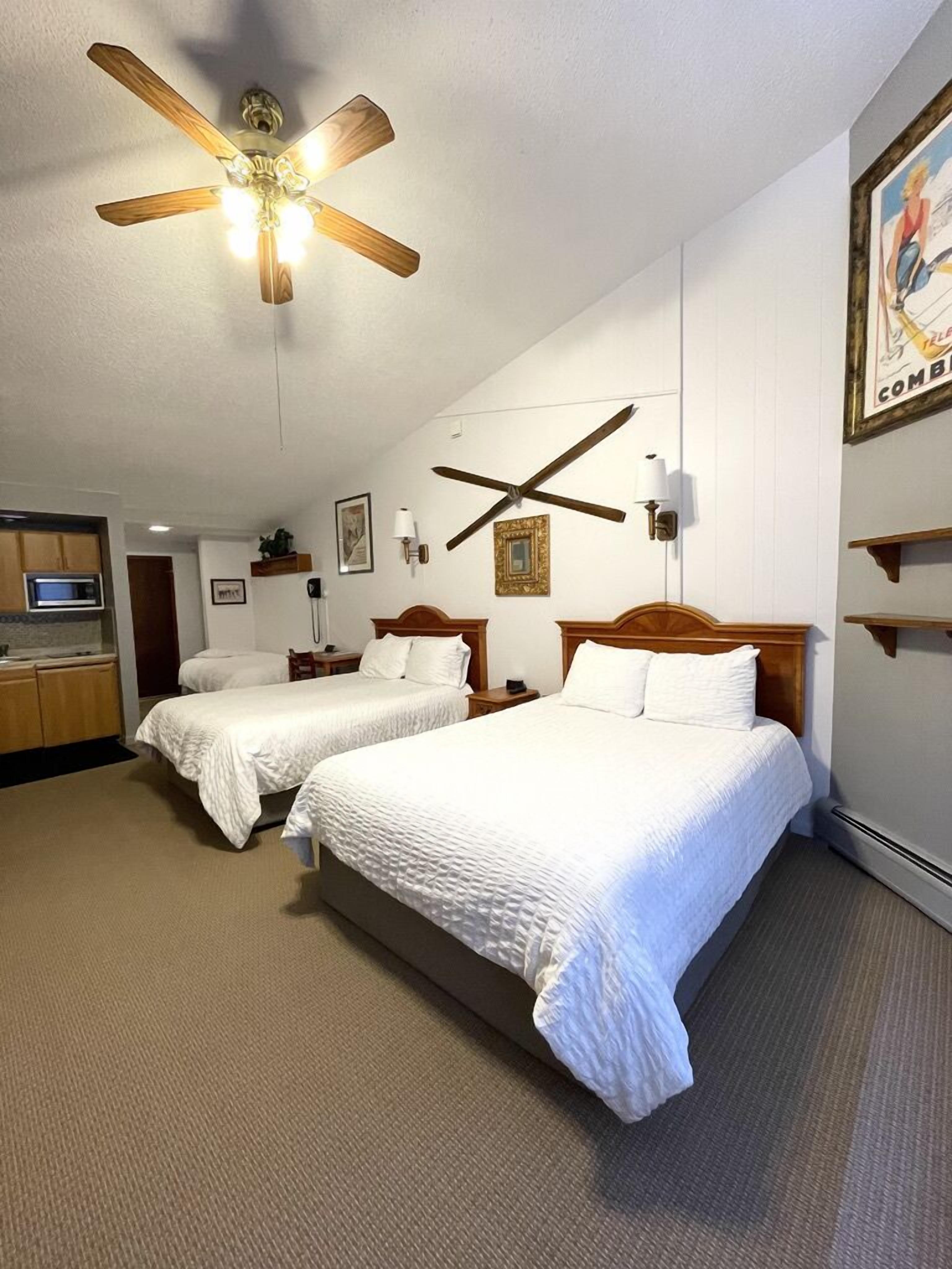A hotel room with two beds, a ceiling fan, a kitchenette, and a painting on the wall.