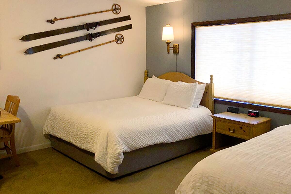 A hotel room with a bed, a nightstand with a lamp, a clock, and skis mounted on the wall.
