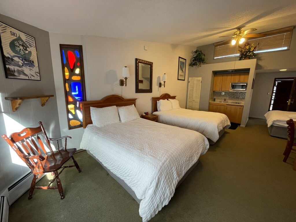 A hotel room with two beds, a wooden chair, a small table, a mirror, and a kitchenette.