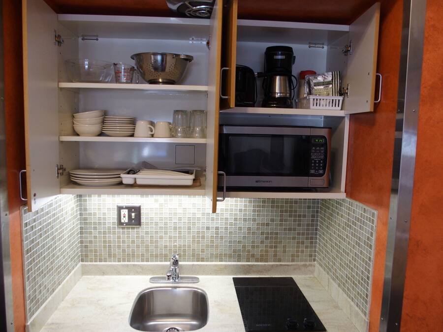 A compact kitchen with a sink, a microwave, and shelves containing dishes and glasses.