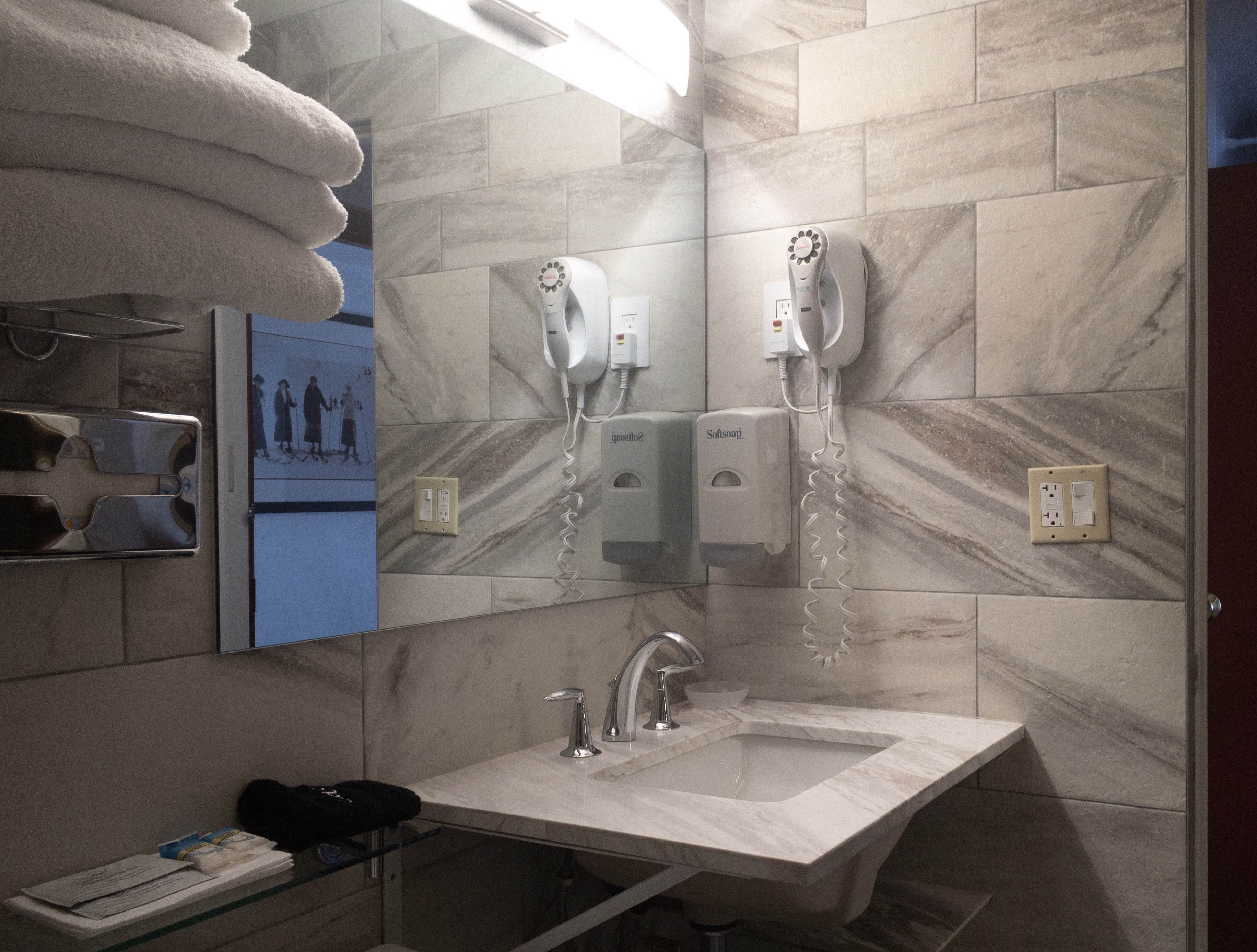 A bathroom with a marble wall, a sink, a mirror, and two hair dryers mounted on the wall.