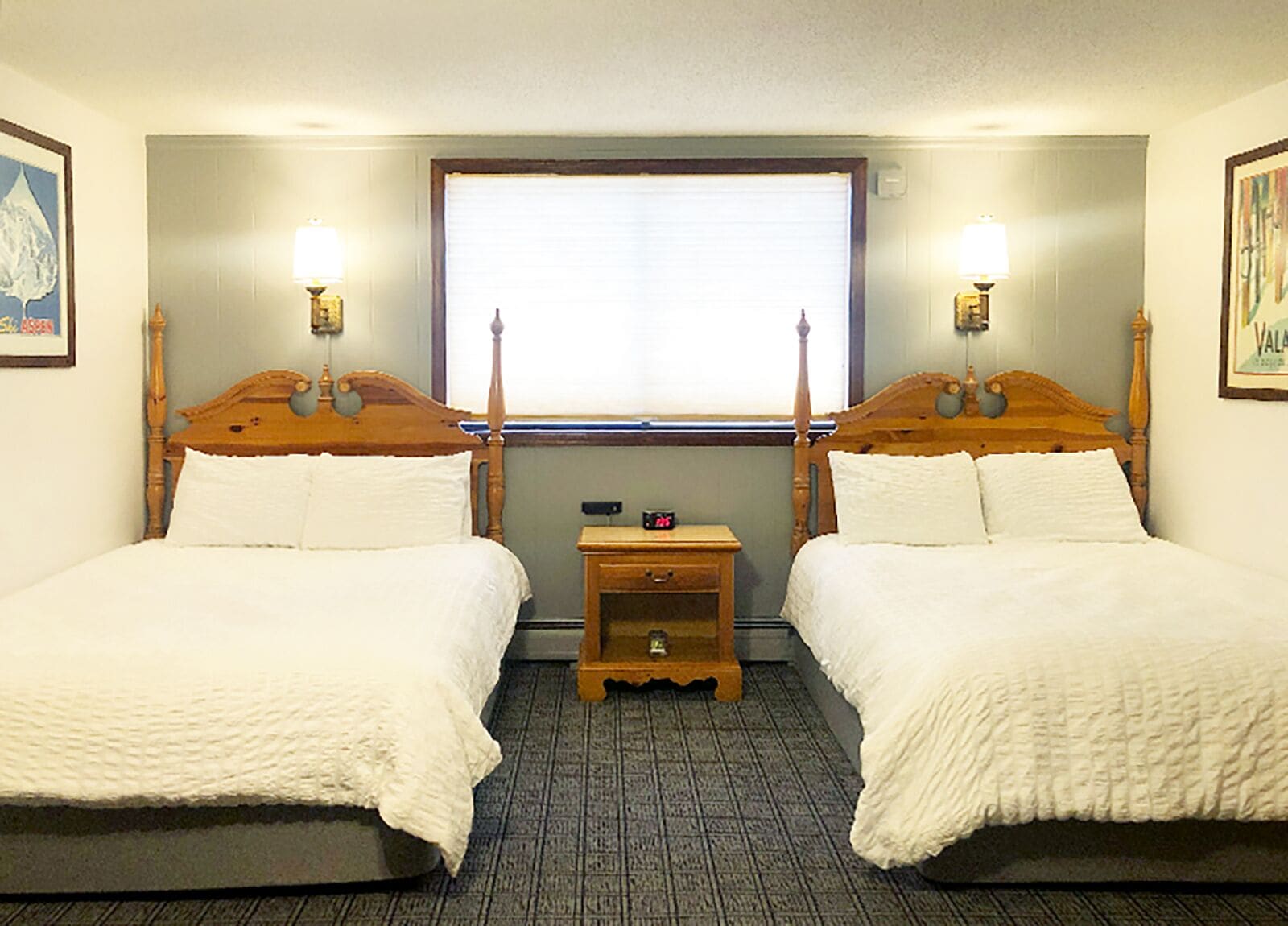 A hotel room with two wooden beds, a small wooden nightstand, a window with blinds, and wall-mounted lamps.