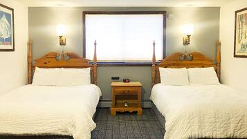 A hotel room with two wooden beds, a small wooden nightstand, a window with blinds, and wall-mounted lamps.