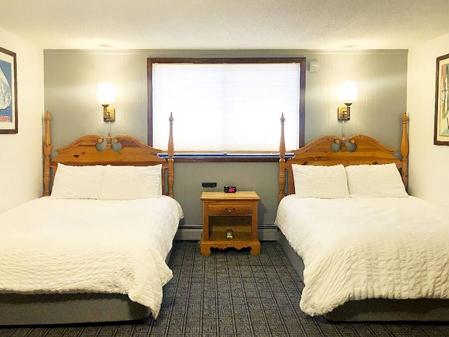 A hotel room with two wooden beds, a small wooden nightstand, a window with blinds, and wall-mounted lamps.