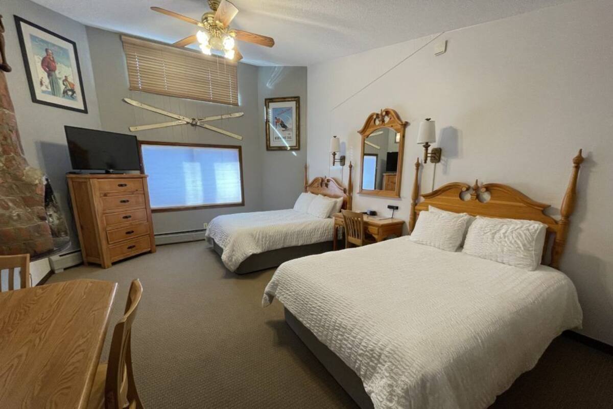 A hotel room with two beds, a wooden desk, a television, and a ceiling fan.