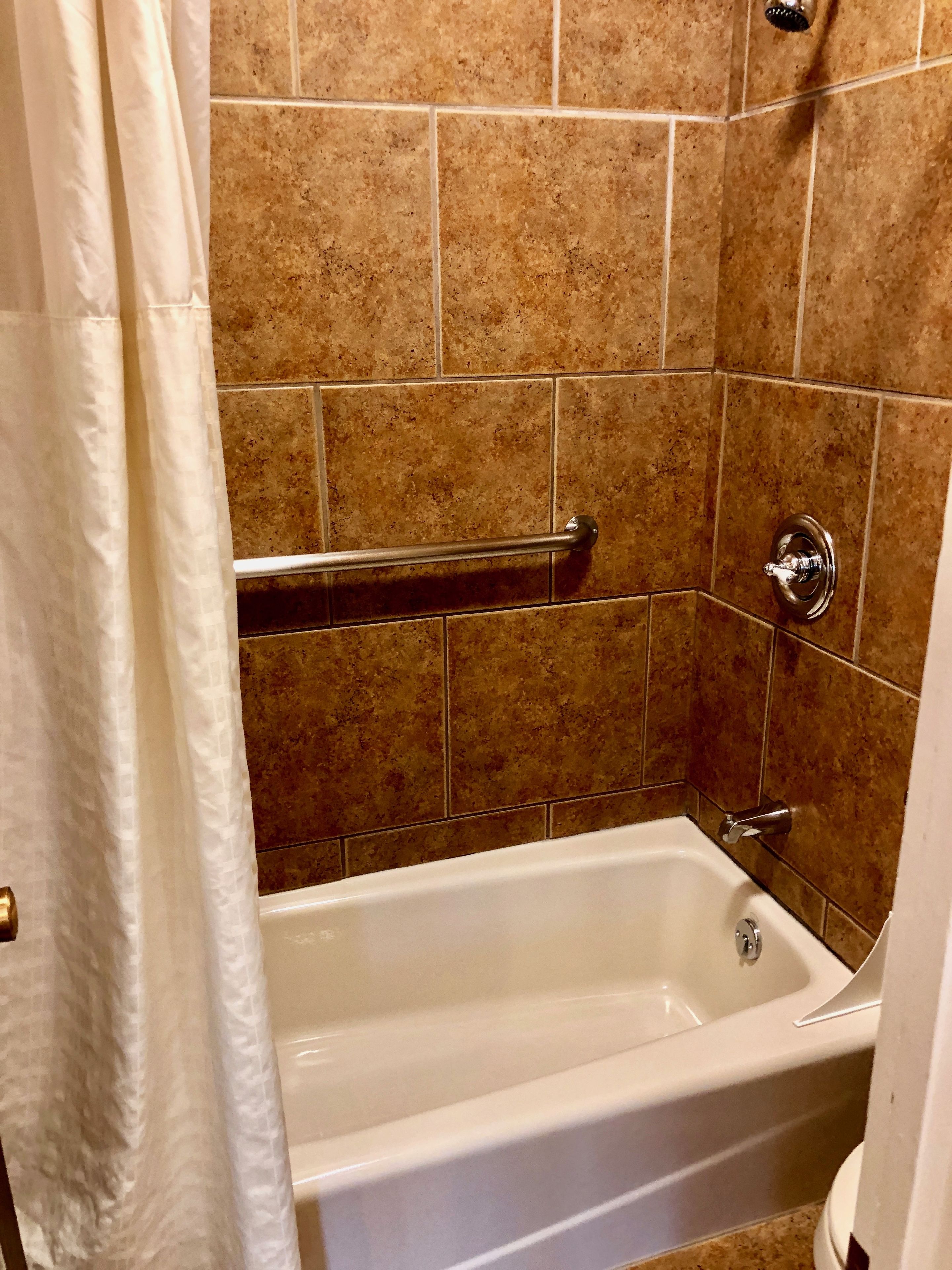 A bathroom with a bathtub, a handrail, and brown tiled walls.