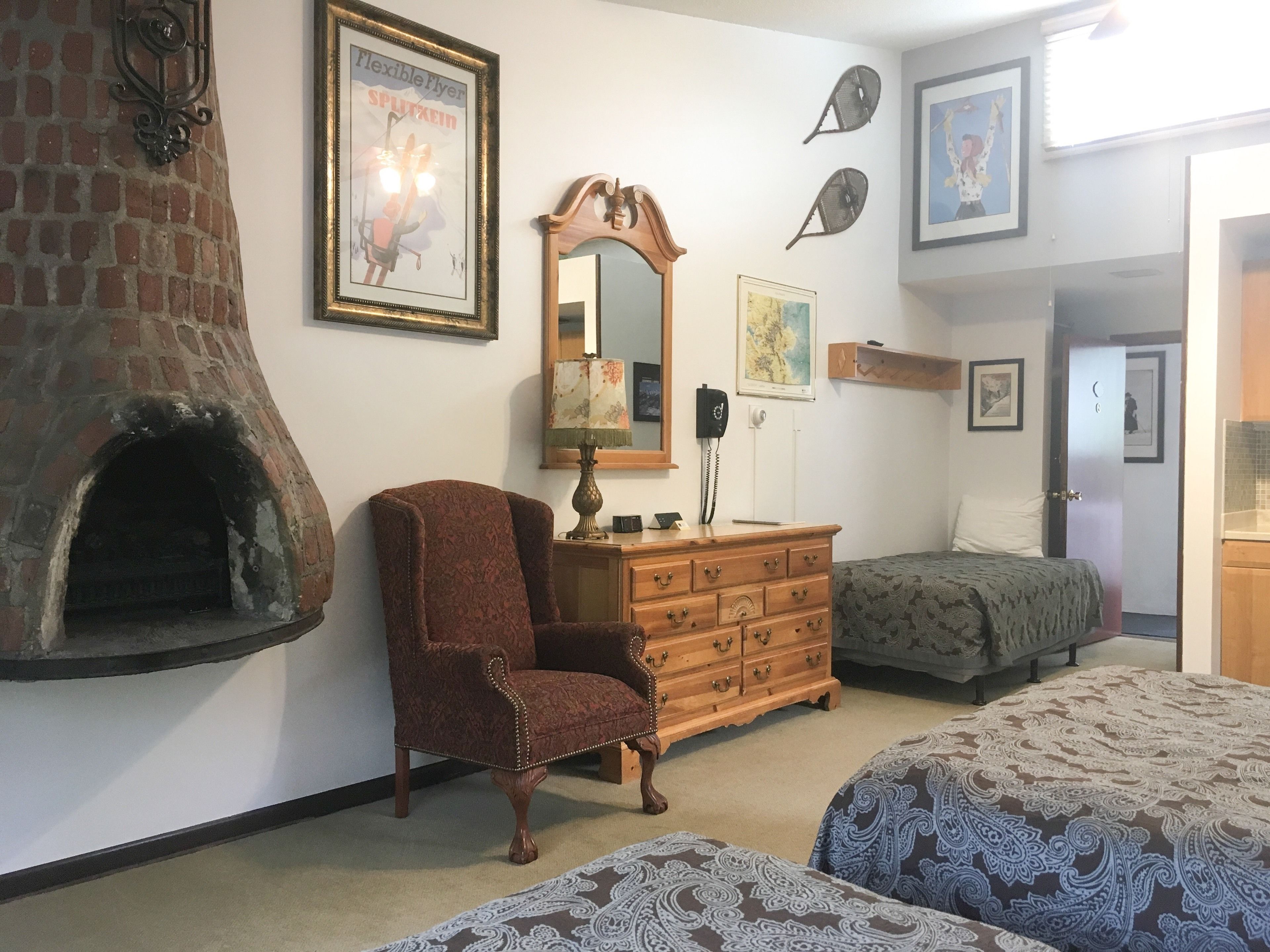 Ein Zimmer mit Kamin aus Ziegelsteinen, einem Holzschrank, einem Stuhl, zwei Betten mit gemusterten Laken sowie verschiedenen Wanddekorationen, darunter ein Spiegel und gerahmte Bilder.