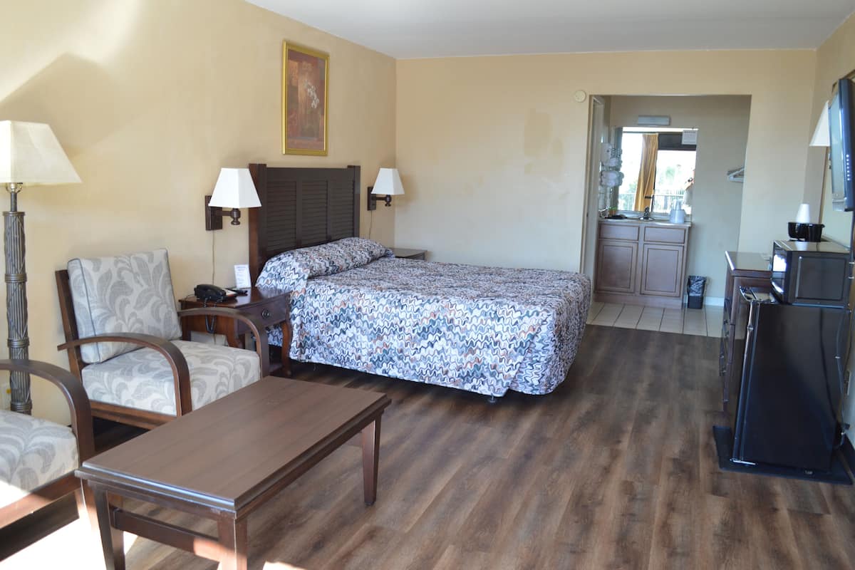 A hotel room with a bed, a desk, a chair, a lamp, a TV, and a kitchenette.