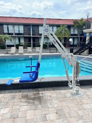 Piscina com cadeira de salva-vidas, placa indicando 'proibido mergulhar' e um edifício com telhado vermelho ao fundo.