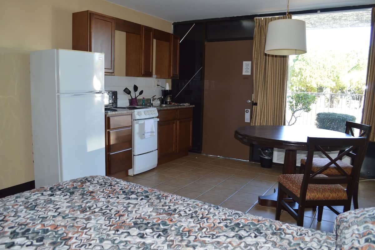A compact kitchen with a refrigerator, stove, and wooden cabinets. There's a dining table with chairs and a patterned bed in the foreground.