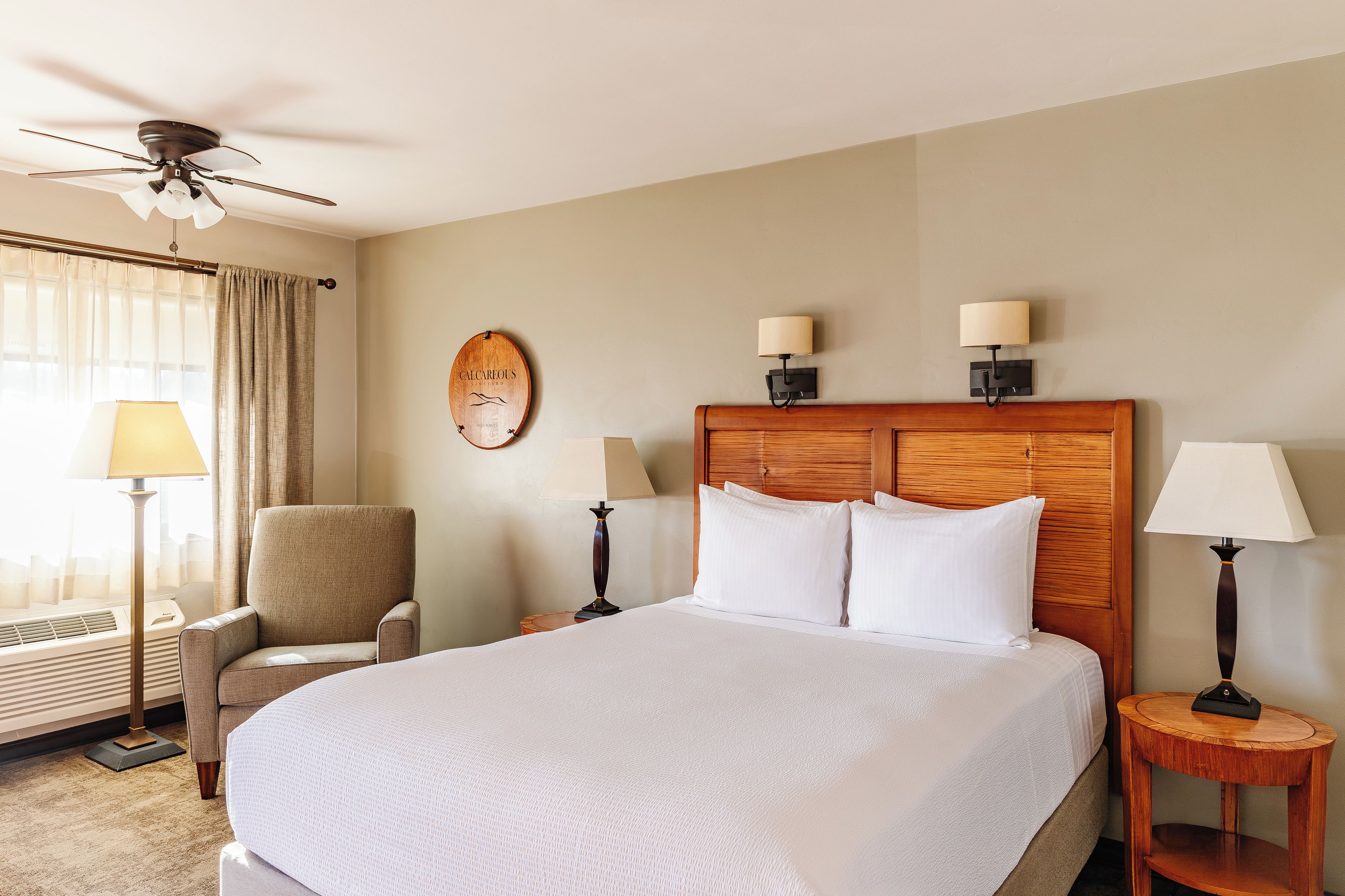A hotel room with a large bed, a chair, a desk, a wall clock, and a ceiling fan.