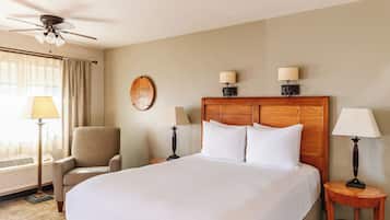 A hotel room with a large bed, a chair, a desk, a wall clock, and a ceiling fan.