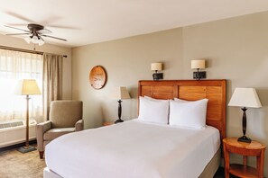 A hotel room with a large bed, a chair, a desk, a wall clock, and a ceiling fan.