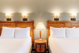 Quarto de hotel com duas camas, uma mesa de cabeceira redonda de madeira e luminárias fixadas na parede.
