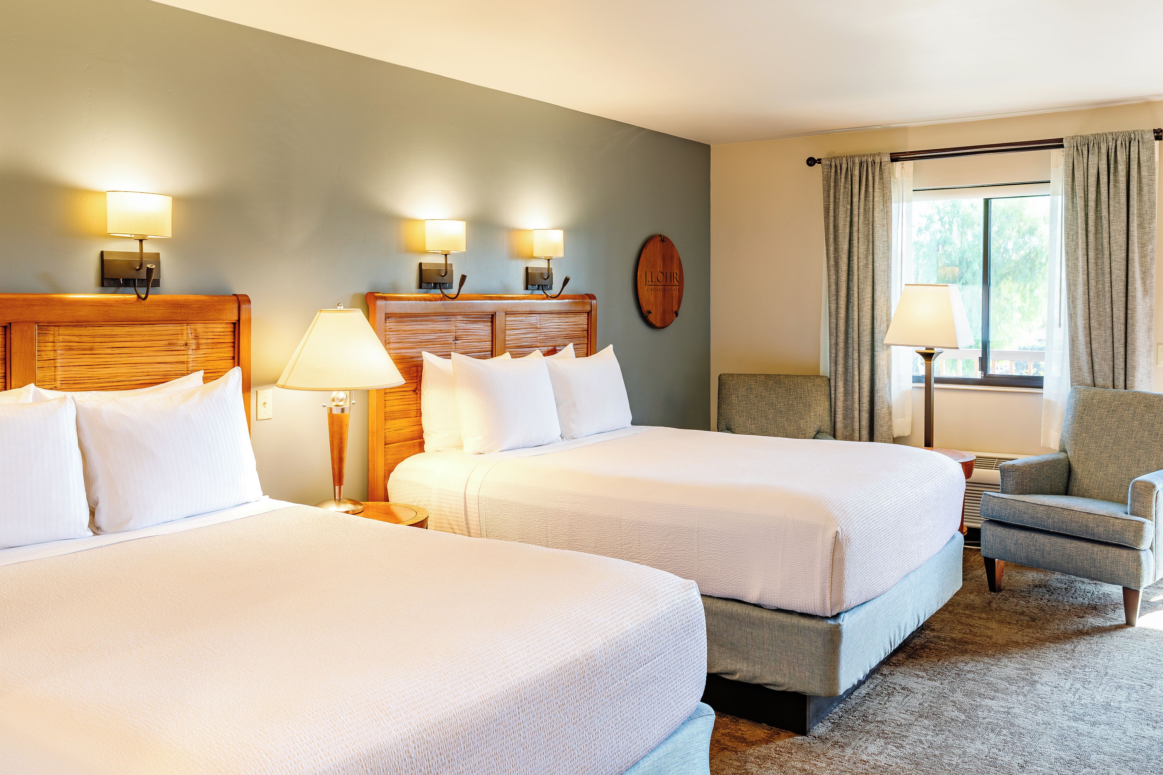A hotel room with two beds, a wooden headboard, a nightstand, a lamp, a chair, and a window with curtains.
